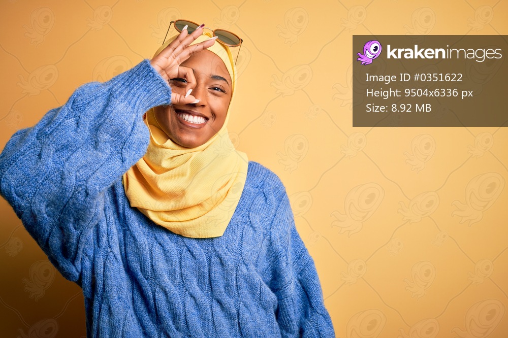 Young African American student woman wearing yellow muslim hijab and sunglasses doing ok gesture with hand smiling, eye looking through fingers with happy face.