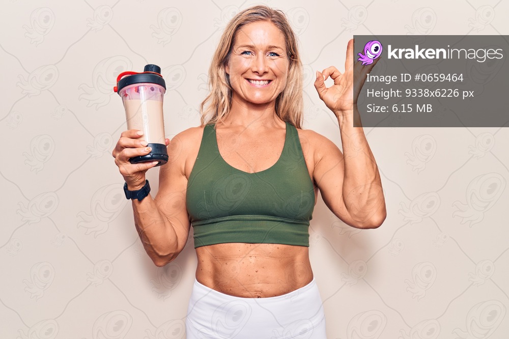 Middle age caucasian blonde woman wearing sport clothes drinking a protein shake doing ok sign with fingers, smiling friendly gesturing excellent symbol 