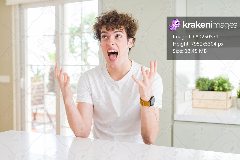 Young handsome man wearing white t-shirt at home crazy and mad shouting and yelling with aggressive expression and arms raised. Frustration concept.