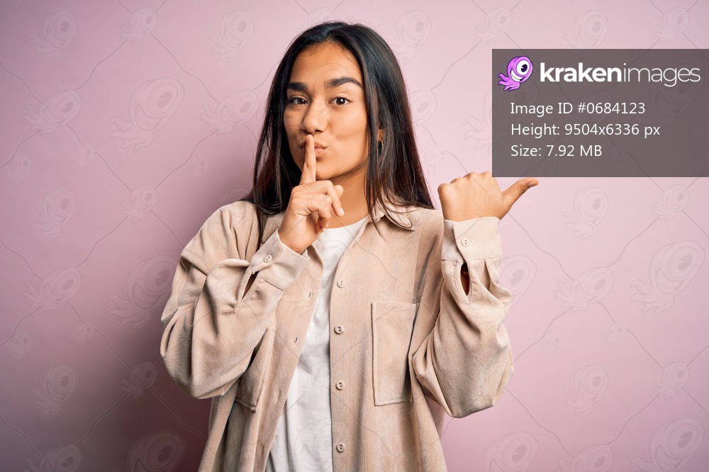 Young beautiful asian woman wearing casual shirt standing over pink background asking to be quiet with finger on lips pointing with hand to the side. Silence and secret concept.