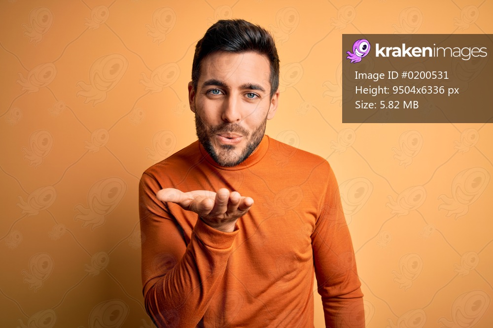 Young handsome man with beard wearing casual sweater standing over yellow background looking at the camera blowing a kiss with hand on air being lovely and sexy. Love expression.