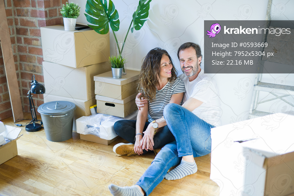 Middle age senior romantic couple in love sitting on the apartment floor with cardboard boxes around and smiling happy for moving to a new home