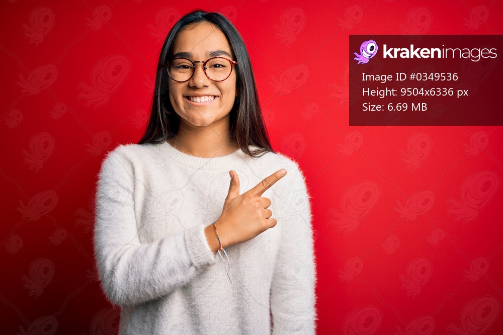 Young beautiful asian woman wearing casual sweater and glasses over red background cheerful with a smile on face pointing with hand and finger up to the side with happy and natural expression