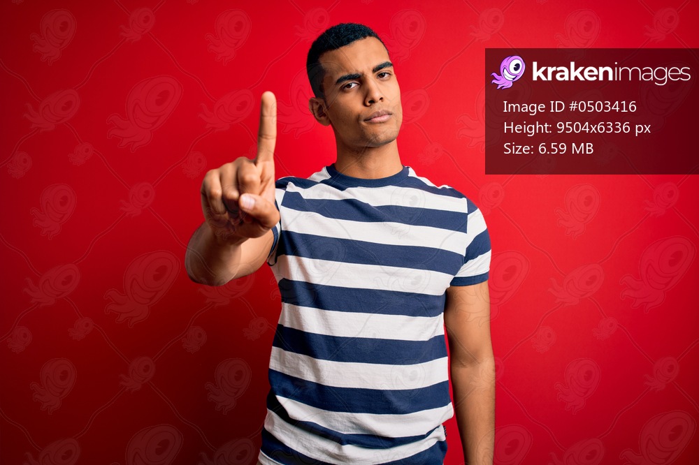 Handsome african american man wearing casual striped t-shirt standing over red background Pointing with finger up and angry expression, showing no gesture