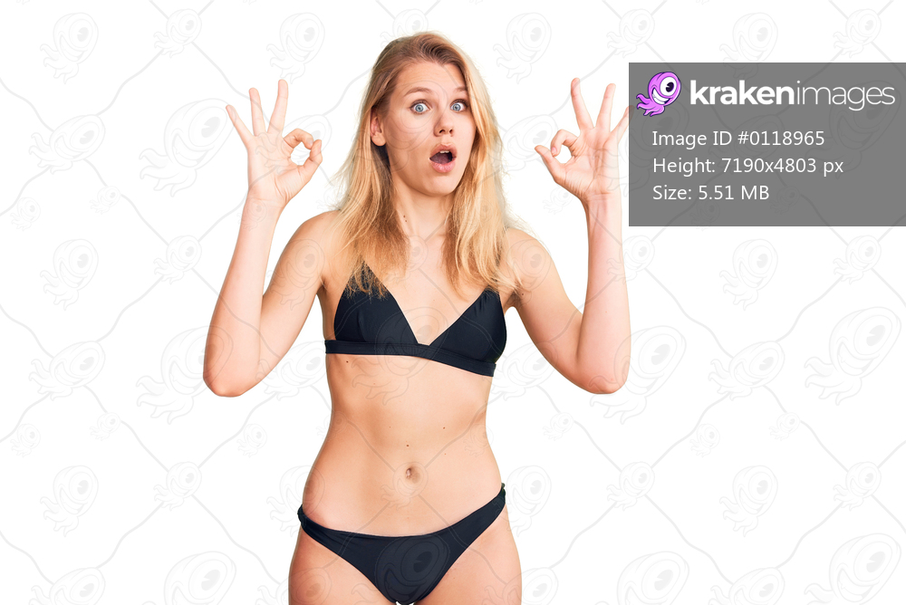 Young beautiful blonde woman wearing bikini looking surprised and shocked doing ok approval symbol with fingers. crazy expression 
