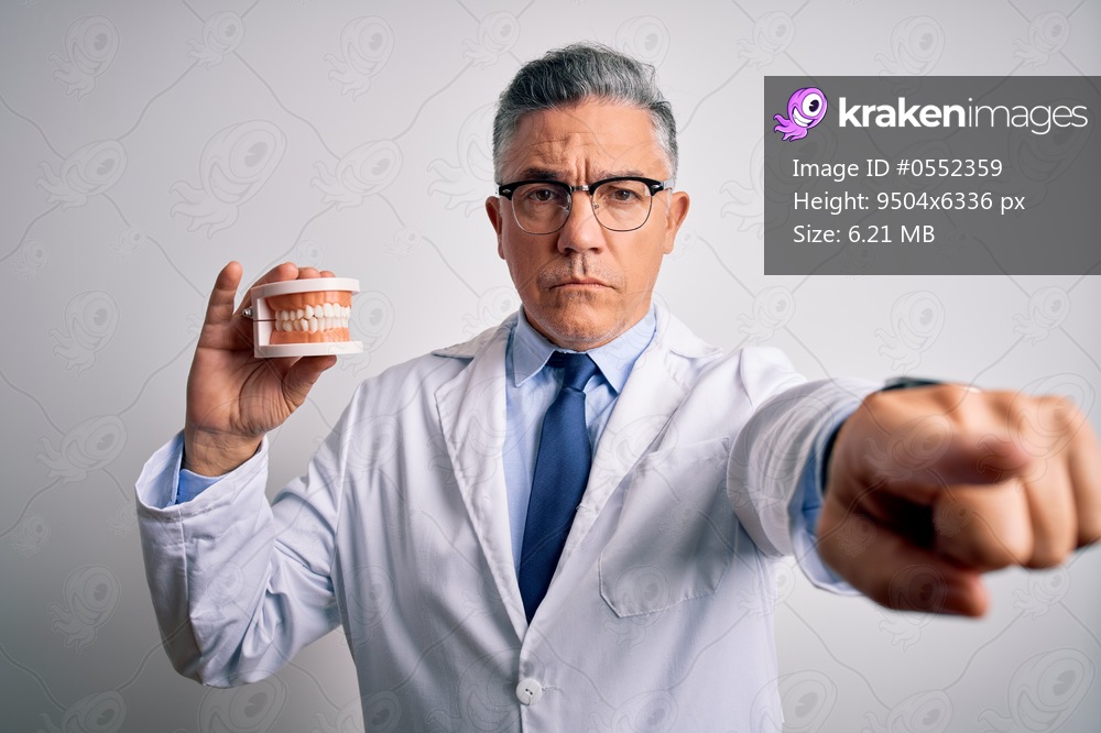 Middle age handsome grey-haired dentist man holding plastic teeth over white background pointing with finger to the camera and to you, hand sign, positive and confident gesture from the front