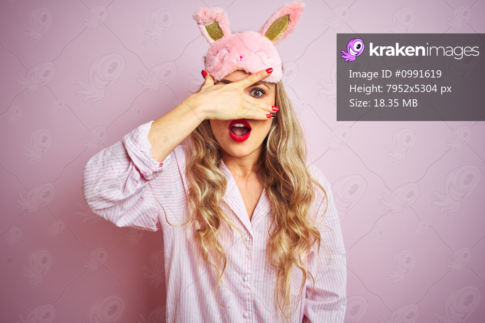 Young beautiful woman wearing pajama and sleep mask over pink isolated background peeking in shock covering face and eyes with hand, looking through fingers with embarrassed expression.