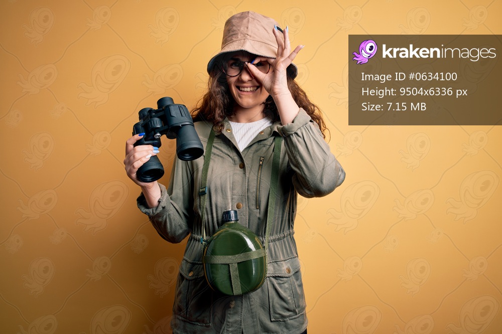 Beautiful tourist woman on vacation wearing explorer hat and water canteen using binoculars with happy face smiling doing ok sign with hand on eye looking through fingers