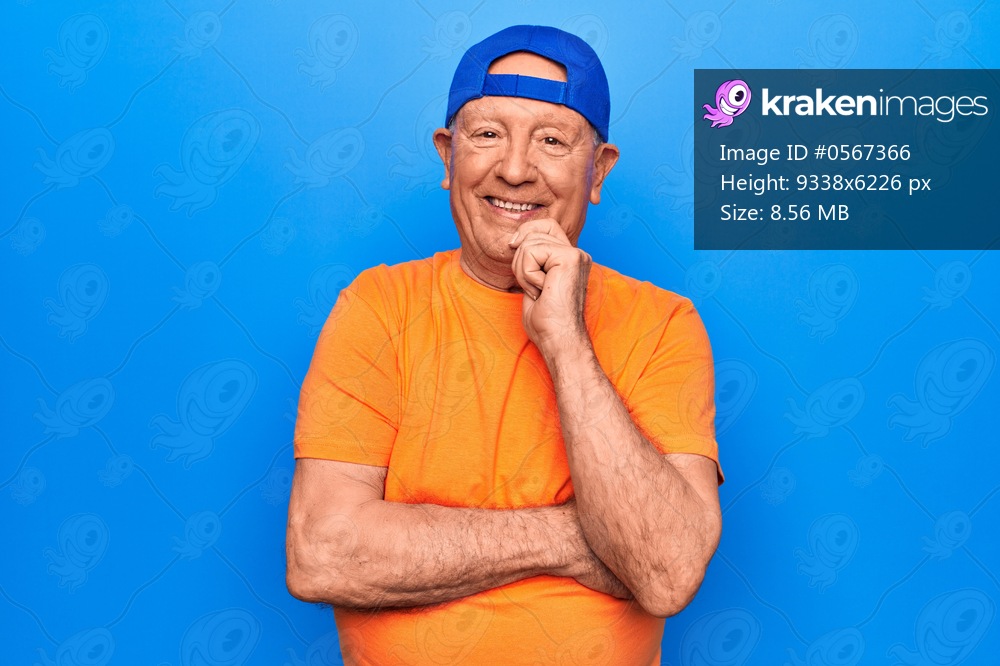 Senior handsome grey-haired man wearing casual t-shirt and cap over blue background smiling looking confident at the camera with crossed arms and hand on chin. Thinking positive.