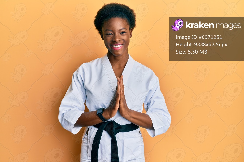 Young african american girl wearing karate kimono and black belt praying with hands together asking for forgiveness smiling confident. 