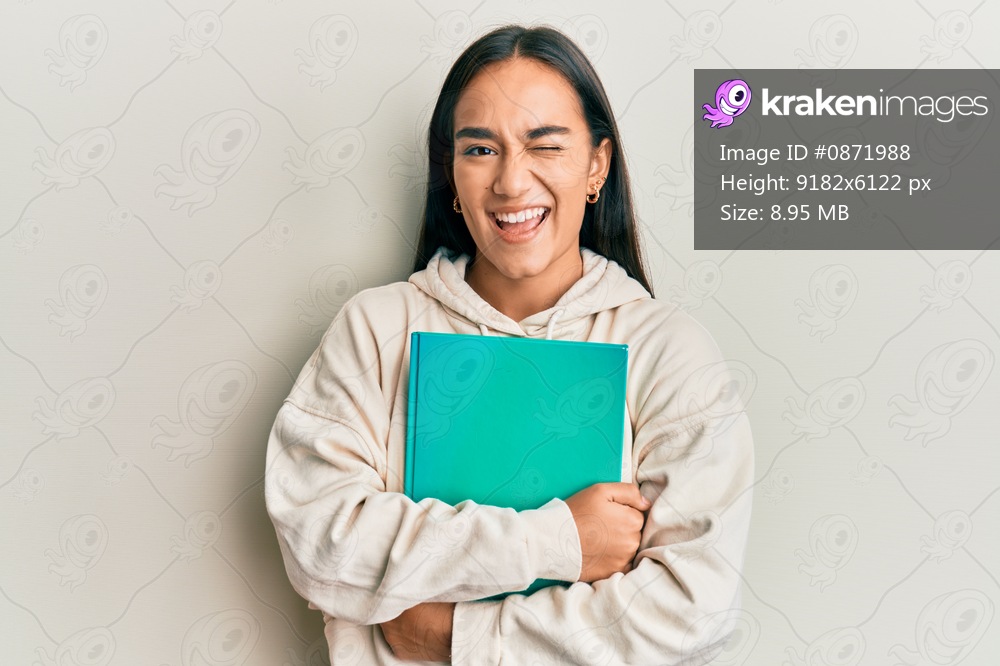 Young asian woman holding book winking looking at the camera with sexy expression, cheerful and happy face. 