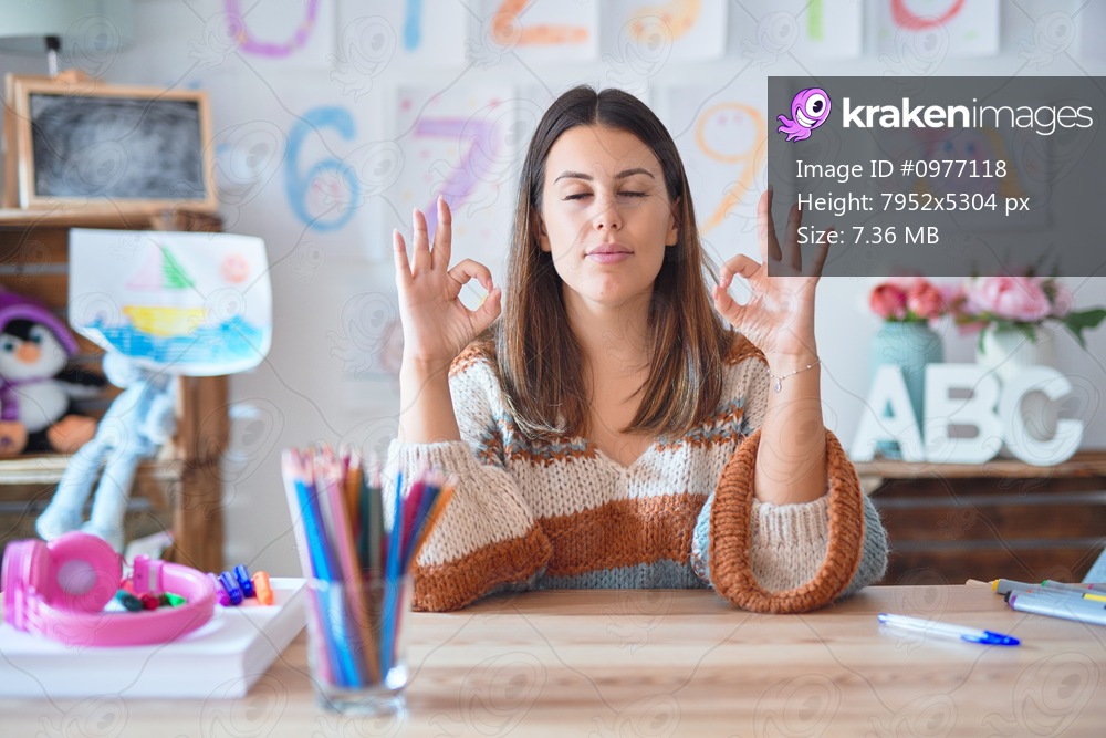 Young beautiful teacher woman wearing sweater and glasses sitting on desk at kindergarten relax and smiling with eyes closed doing meditation gesture with fingers. Yoga concept.