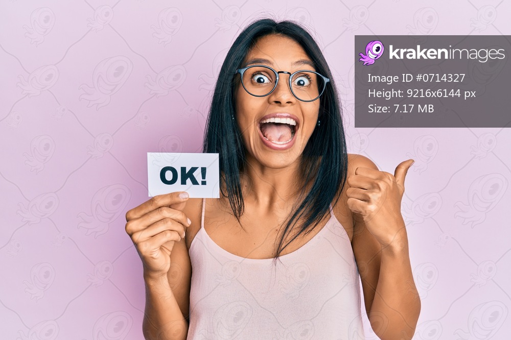 Young african american woman holding ok message paper pointing thumb up to the side smiling happy with open mouth 