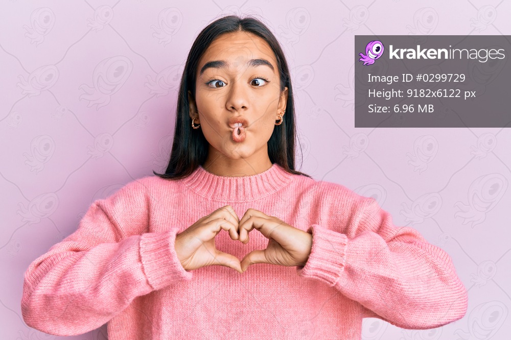 Young asian woman doing heart symbol with hands making fish face with mouth and squinting eyes, crazy and comical. 