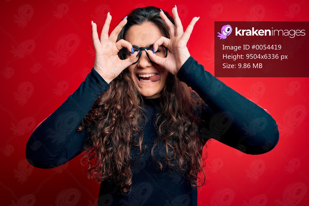 Young beautiful woman with curly hair wearing sweater and glasses over red background doing ok gesture like binoculars sticking tongue out, eyes looking through fingers. Crazy expression.