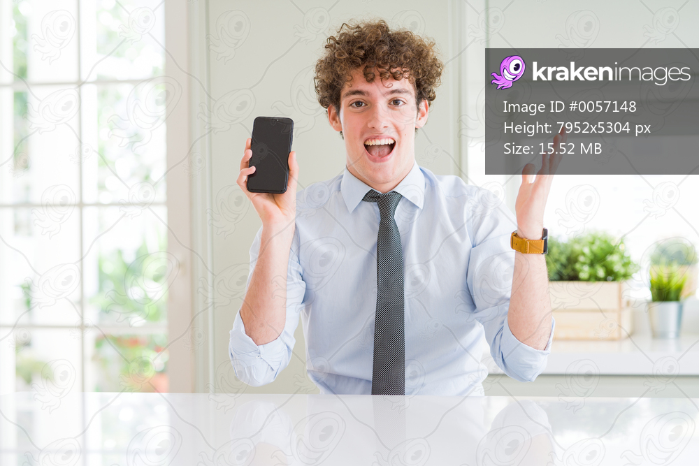 Young business man showing smartphone screen at the office very happy and excited, winner expression celebrating victory screaming with big smile and raised hands