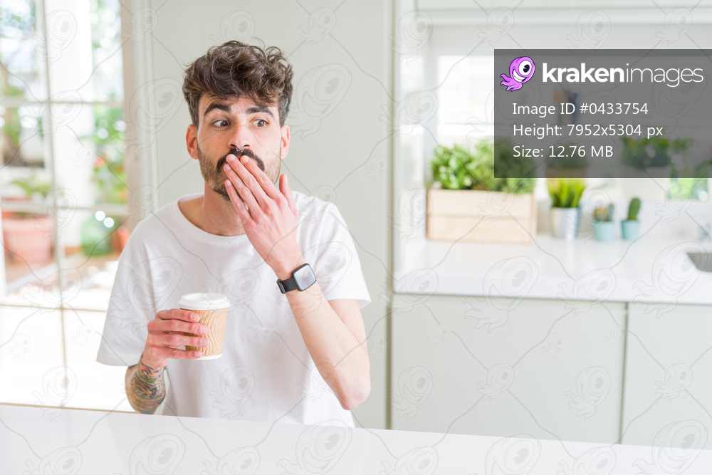 Young man drinking a coffee in a paper cup in the morning cover mouth with hand shocked with shame for mistake, expression of fear, scared in silence, secret concept