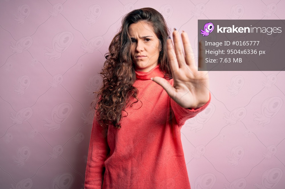 Young beautiful woman with curly hair wearing turtleneck sweater over pink background doing stop sing with palm of the hand. Warning expression with negative and serious gesture on the face.