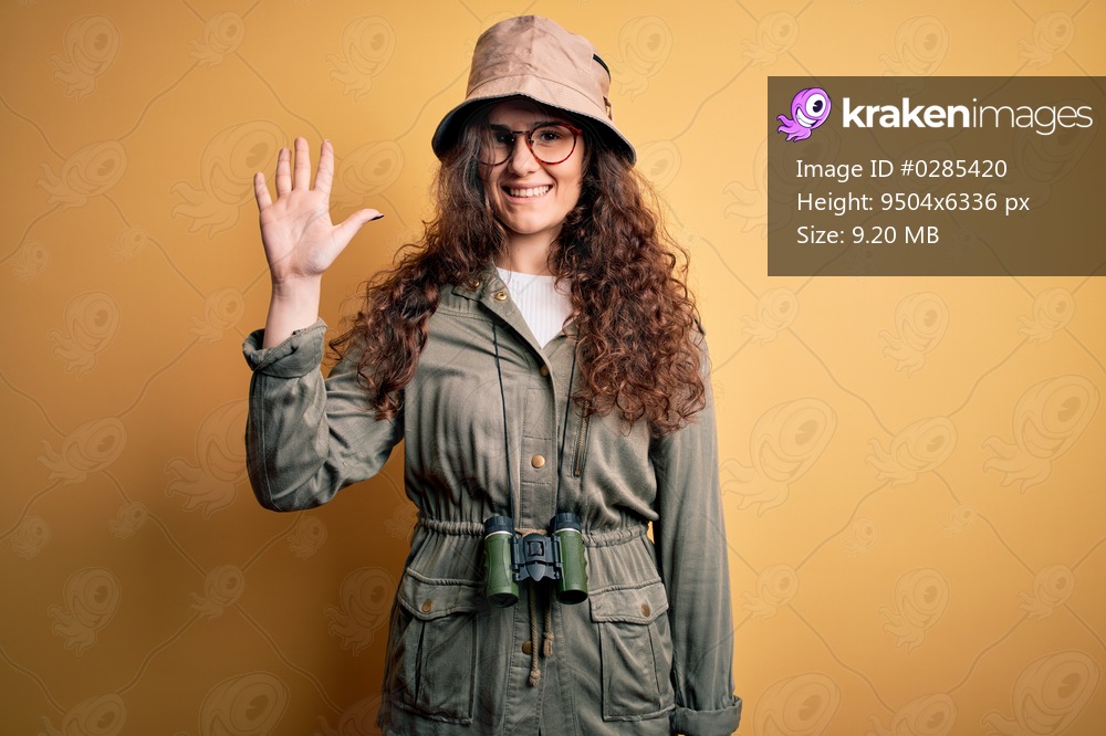 Young beautiful tourist woman on vacation wearing explorer hat and binoculars showing and pointing up with fingers number five while smiling confident and happy.