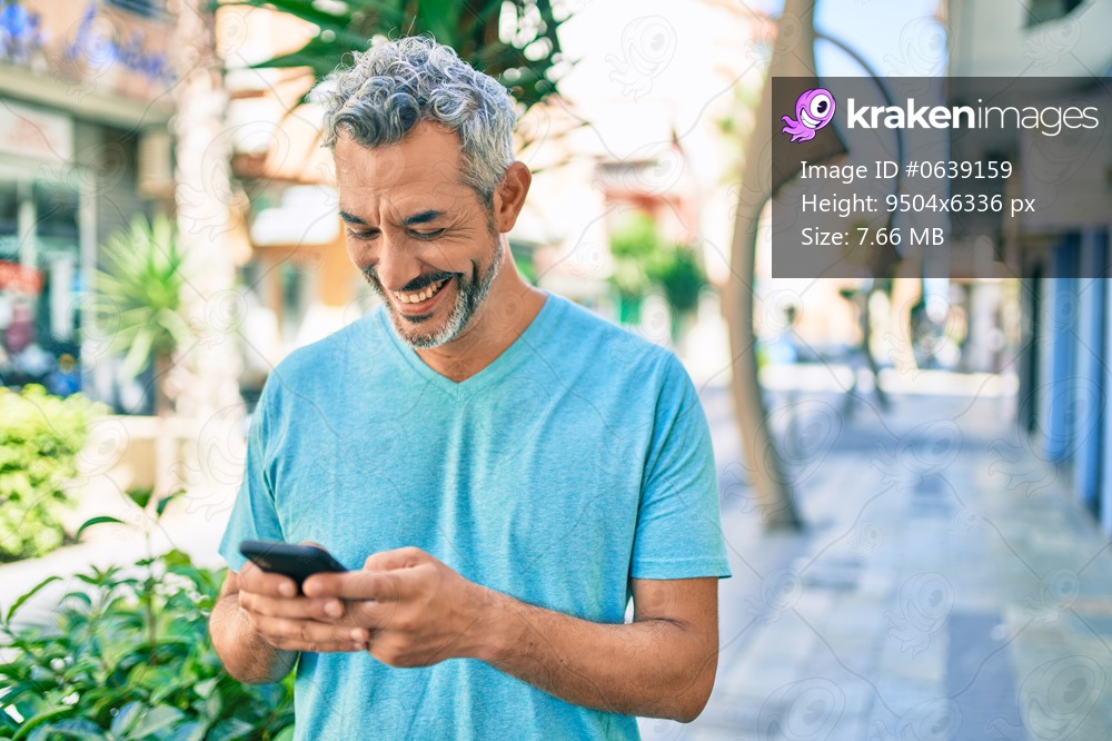 Middle age grey-haired man smiling happy using smartphone walking at street of city.