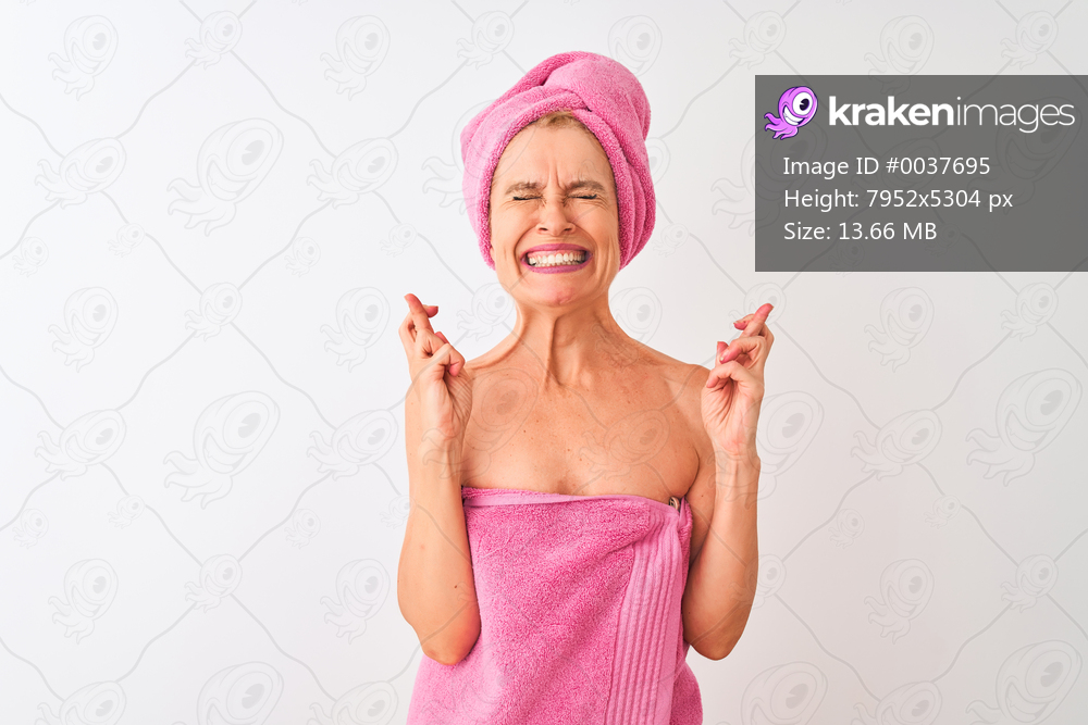 Middle age woman wearing shower towel after bath standing over isolated white background gesturing finger crossed smiling with hope and eyes closed. Luck and superstitious concept.