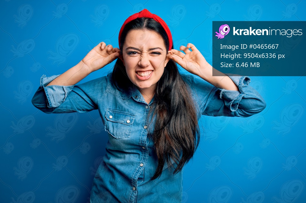 Young brunette woman wearing casual denim shirt over blue isolated background Smiling pulling ears with fingers, funny gesture. Audition problem