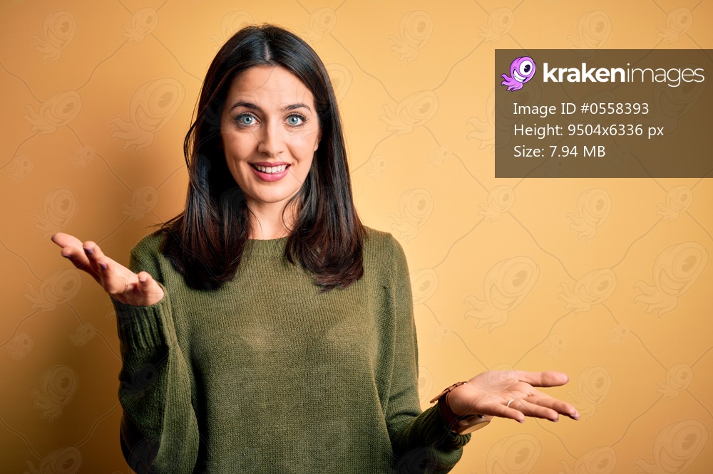 Young brunette woman with blue eyes wearing green casual sweater over yellow background smiling cheerful with open arms as friendly welcome, positive and confident greetings