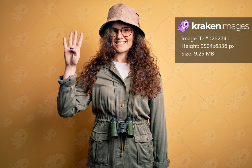 Young beautiful tourist woman on vacation wearing explorer hat and binoculars showing and pointing up with fingers number four while smiling confident and happy.