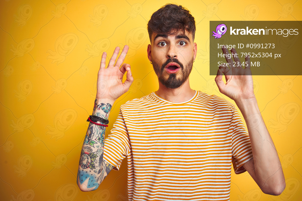 Young man with tattoo wearing striped t-shirt standing over isolated yellow background looking surprised and shocked doing ok approval symbol with fingers. Crazy expression