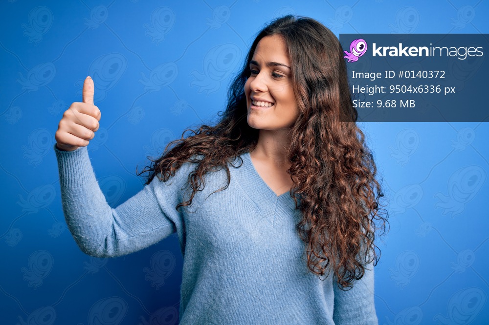 Young beautiful woman with curly hair wearing blue casual sweater over isolated background Looking proud, smiling doing thumbs up gesture to the side