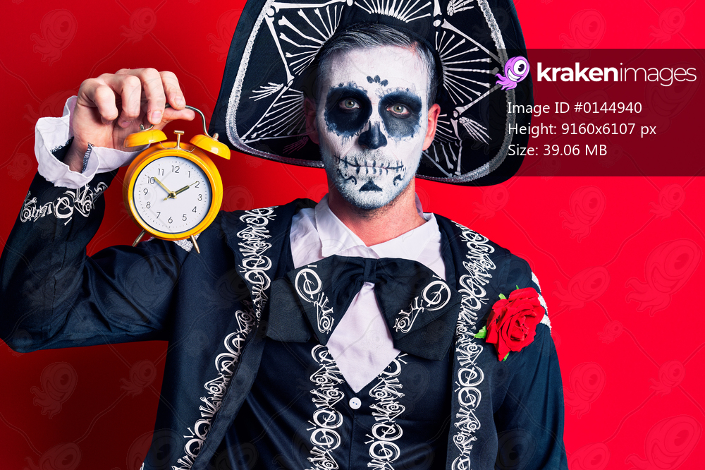 Young man wearing mexican day of the dead costume holding alarm clock thinking attitude and sober expression looking self confident 