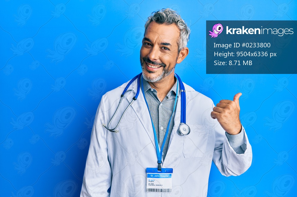 Middle age grey-haired man wearing doctor uniform and stethoscope smiling with happy face looking and pointing to the side with thumb up. 
