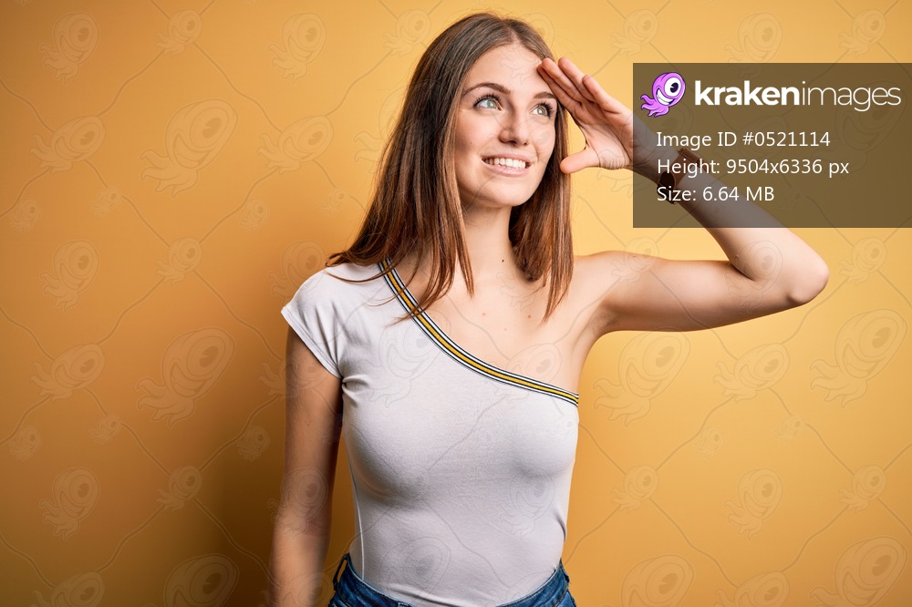 Young beautiful redhead woman wearing casual t-shirt over isolated yellow background very happy and smiling looking far away with hand over head. Searching concept.