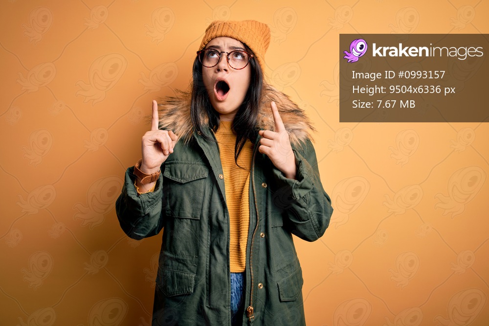 Young brunette woman wearing glasses and winter coat with hat over yellow isolated background amazed and surprised looking up and pointing with fingers and raised arms.