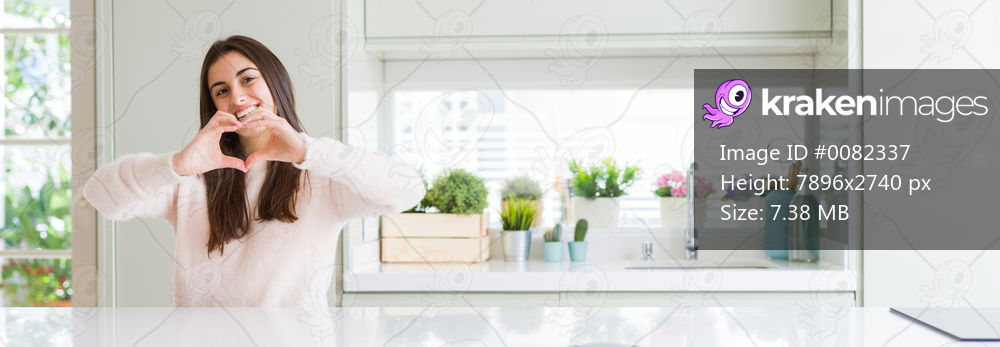 Wide angle picture of beautiful young woman sitting on white table at home smiling in love showing heart symbol and shape with hands. Romantic concept.