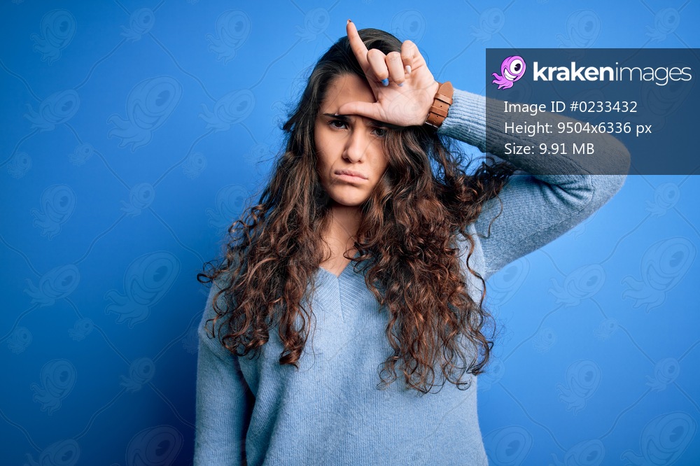 Young beautiful woman with curly hair wearing blue casual sweater over isolated background making fun of people with fingers on forehead doing loser gesture mocking and insulting.