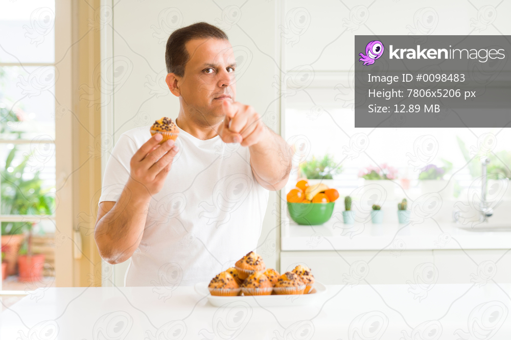 Middle age man eating chocolate chip muffins at home pointing with finger to the camera and to you, hand sign, positive and confident gesture from the front