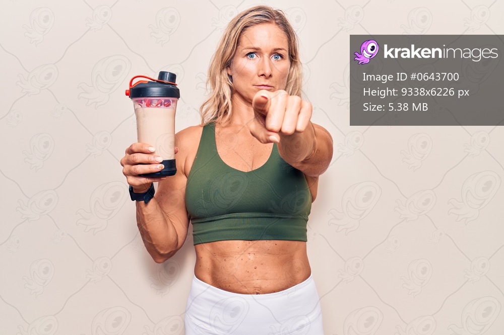 Middle age caucasian blonde woman wearing sport clothes drinking a protein shake pointing with finger to the camera and to you, confident gesture looking serious 