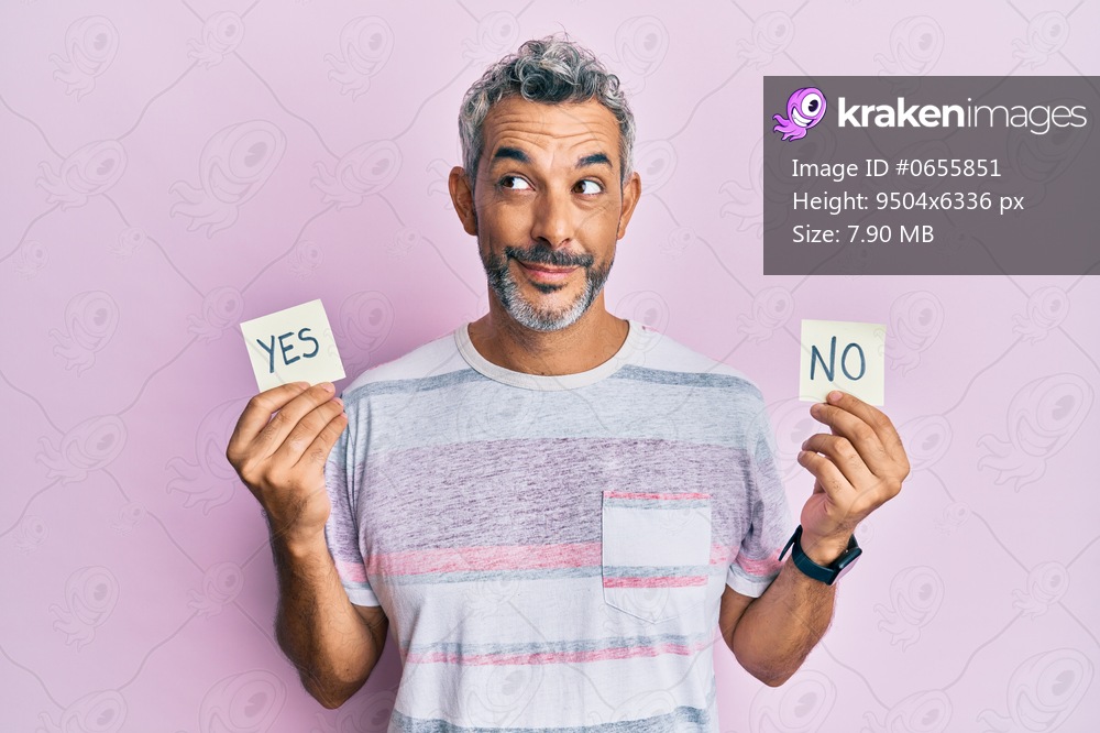 Middle age grey-haired man holding yes and no reminder smiling looking to the side and staring away thinking. 