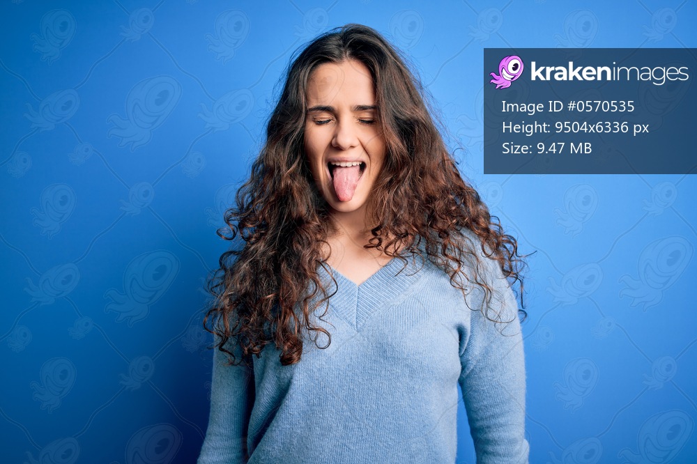 Young beautiful woman with curly hair wearing blue casual sweater over isolated background sticking tongue out happy with funny expression. Emotion concept.