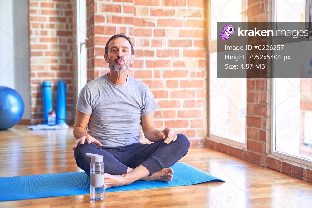 Middle age handsome sportman sitting on mat doing stretching yoga exercise at gym looking at the camera blowing a kiss on air being lovely and sexy. Love expression.