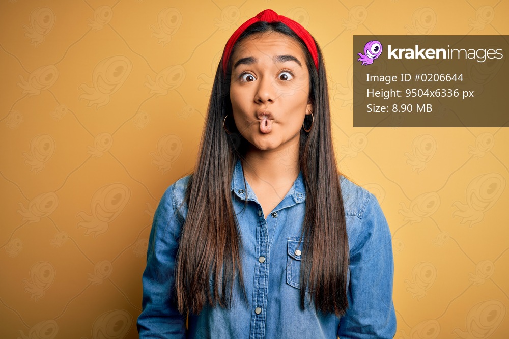 Young beautiful asian woman wearing casual denim shirt and diadem over yellow background making fish face with lips, crazy and comical gesture. Funny expression.