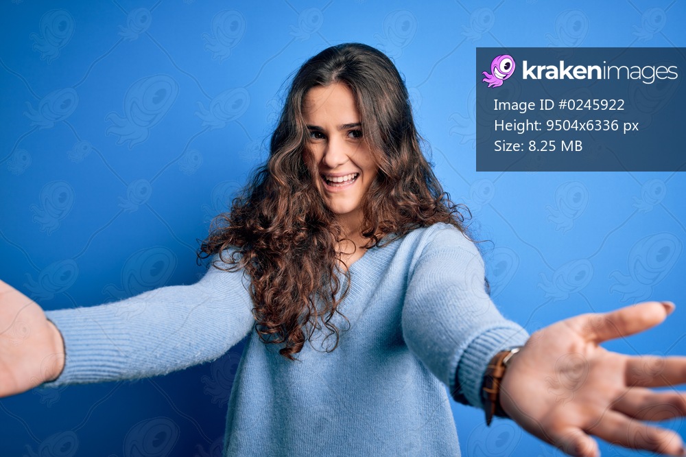 Young beautiful woman with curly hair wearing blue casual sweater over isolated background looking at the camera smiling with open arms for hug. Cheerful expression embracing happiness.