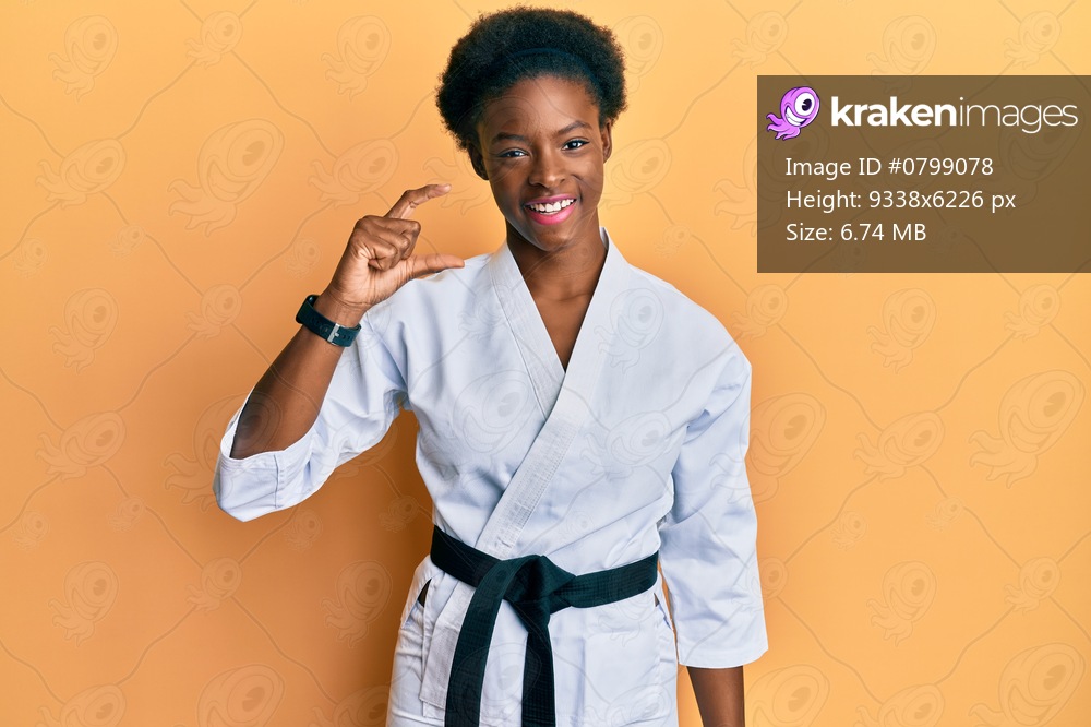 Young african american girl wearing karate kimono and black belt smiling and confident gesturing with hand doing small size sign with fingers looking and the camera. measure concept. 