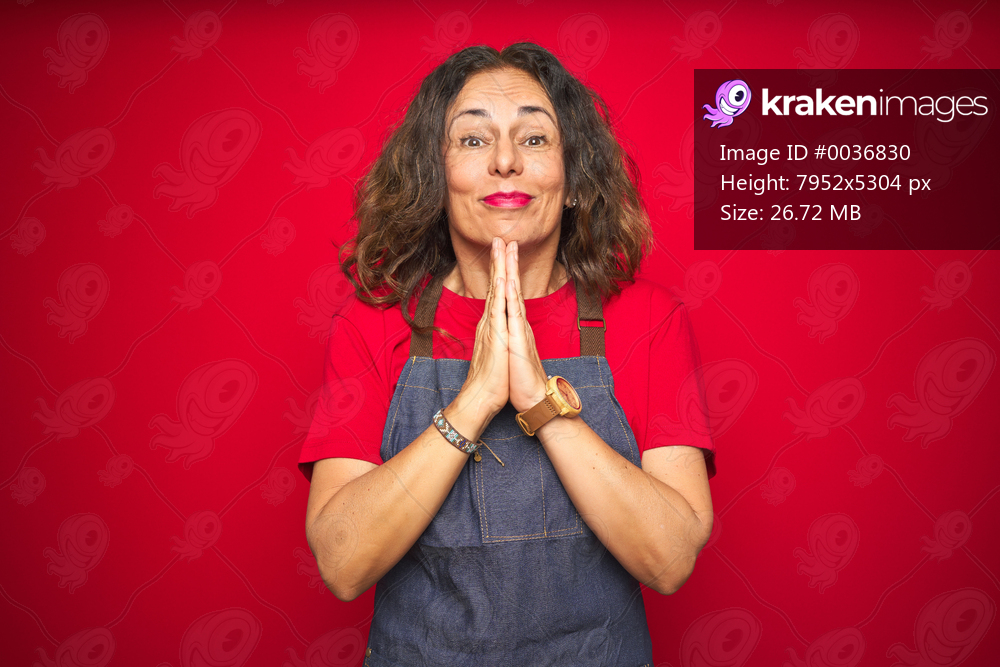 Middle age senior woman wearing apron uniform over red isolated background praying with hands together asking for forgiveness smiling confident.