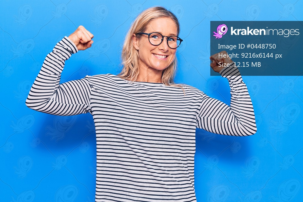 Middle age caucasian blonde woman wearing casual striped sweater and glasses showing arms muscles smiling proud. fitness concept. 