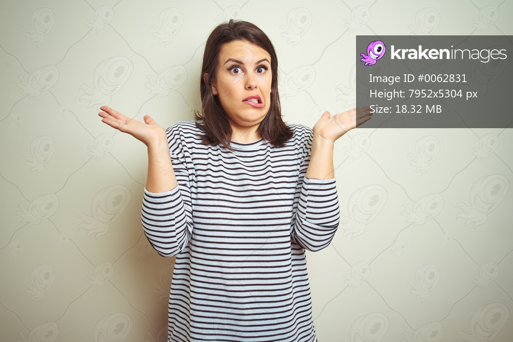 Young beautiful brunette woman wearing striped sweater over white isolated background clueless and confused expression with arms and hands raised. Doubt concept.