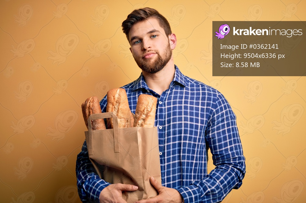 Young blond man with beard and blue eyes holding paper bag with bread with a confident expression on smart face thinking serious