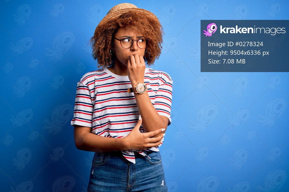 African American woman with curly hair on vacation wearing summer hat and striped t-shirt looking stressed and nervous with hands on mouth biting nails. Anxiety problem.