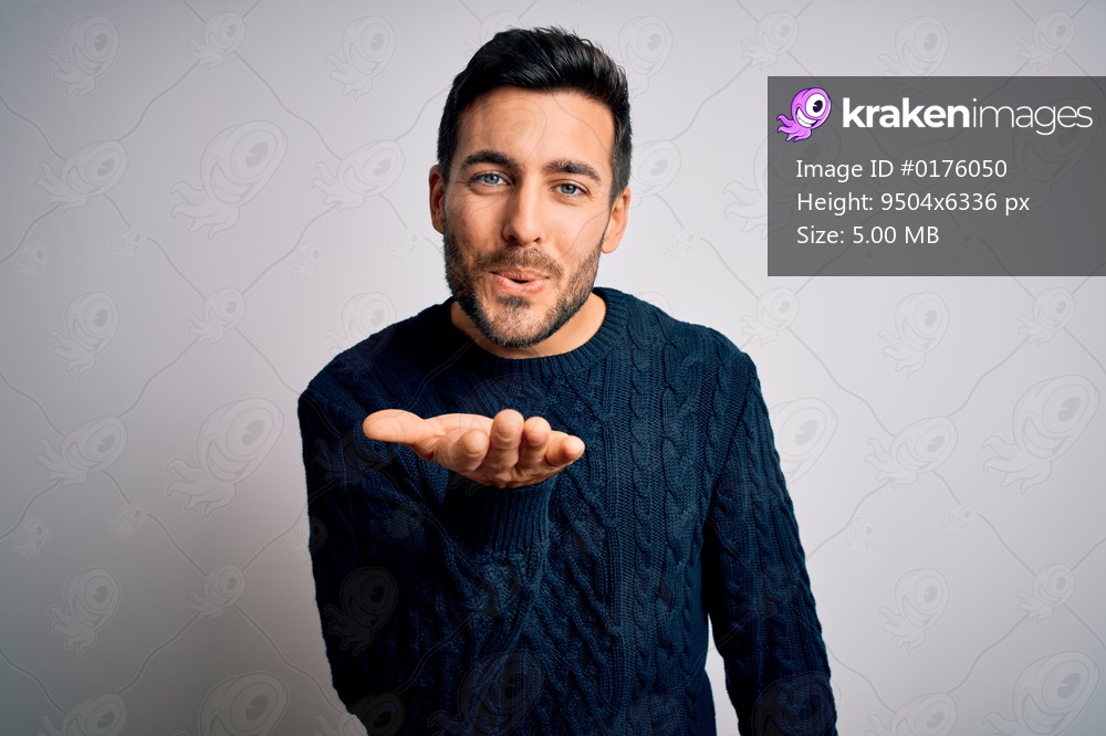 Young handsome man with beard wearing casual sweater standing over white background looking at the camera blowing a kiss with hand on air being lovely and sexy. Love expression.